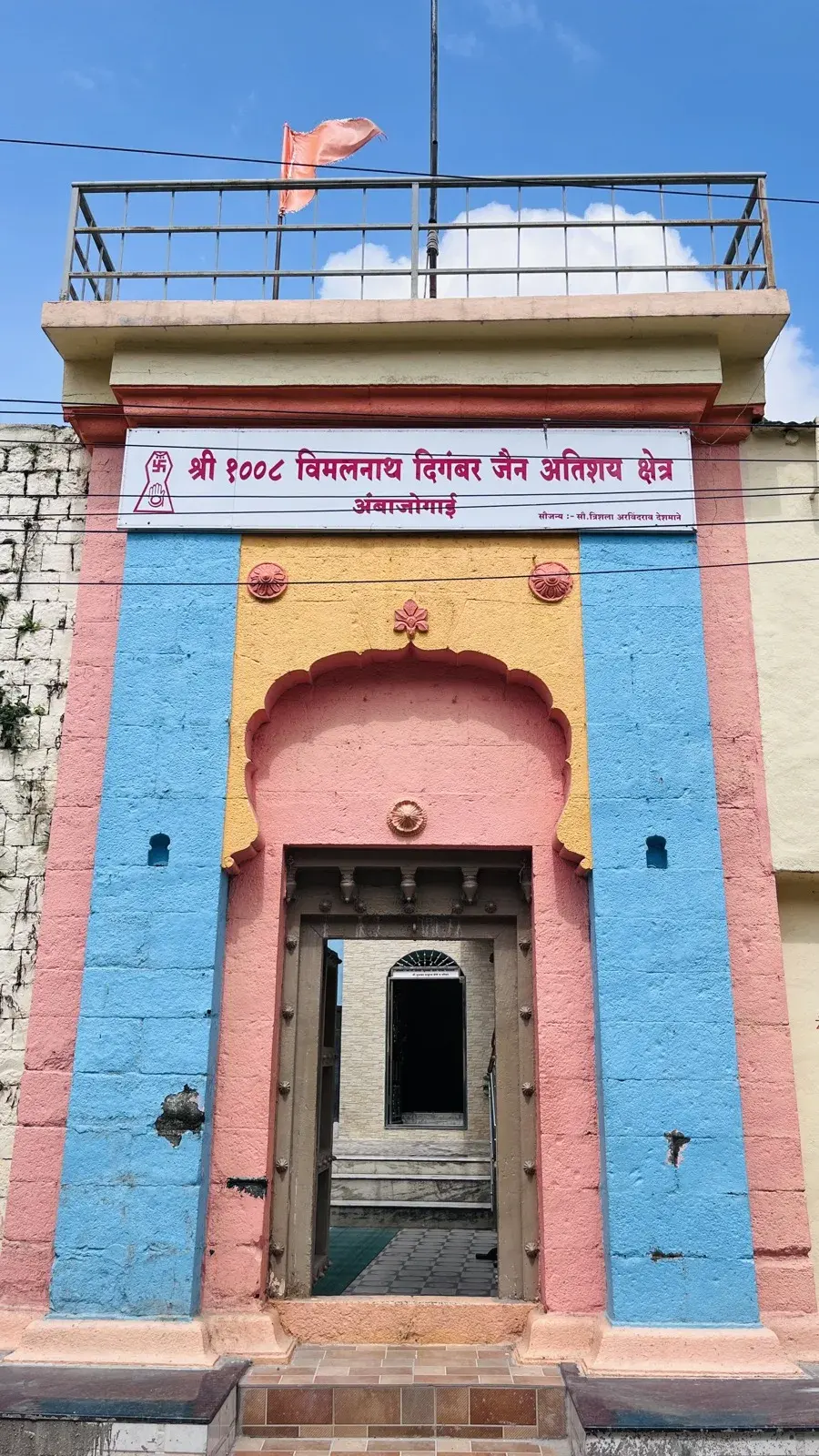 The west-facing gateway of the kshetra from Jain Galli, painted pink, blue and yellow, with the signboard 'Shri 1008 Vimalnath Digambar Jain Atishay Kshetra, Ambajogai' over the entrance and the garbhagriha visible through the open doorway.
