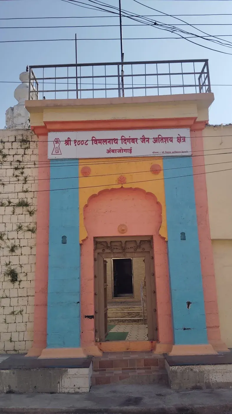 The kshetra's entrance gate on Jain Galli, pink, blue and yellow, its signboard reading 'Shri 1008 Vimalnath Digambar Jain Atishay Kshetra' above the open doorway, a second temple's white shikhar visible beyond the boundary wall.