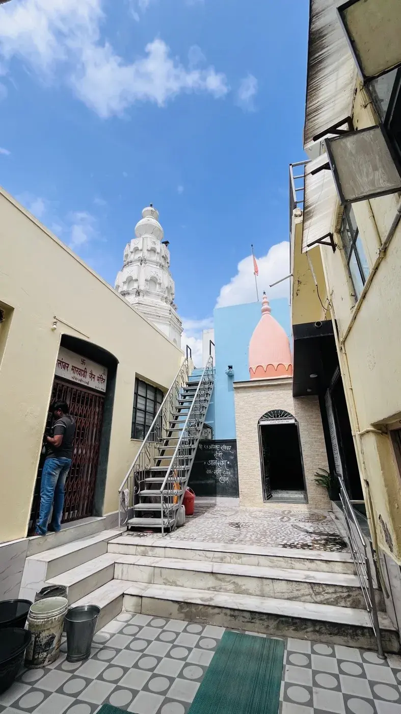 A courtyard within the kshetra complex, a metal staircase leading up past a small pink-domed shrine, with the white shikhar of the neighbouring Shwetambar Marwadi Jain mandir visible beyond the rooftops.