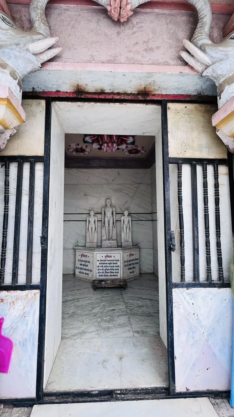 Three standing white marble Tirthankara murtis beneath a hand-painted lotus ceiling, donor names inscribed on the pedestal below.