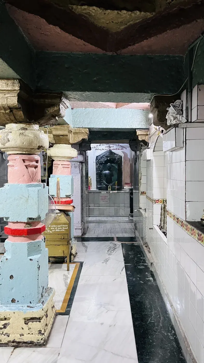 A pastel-pillared corridor within the kshetra leading toward a small black stone shrine glimpsed in the archway beyond.