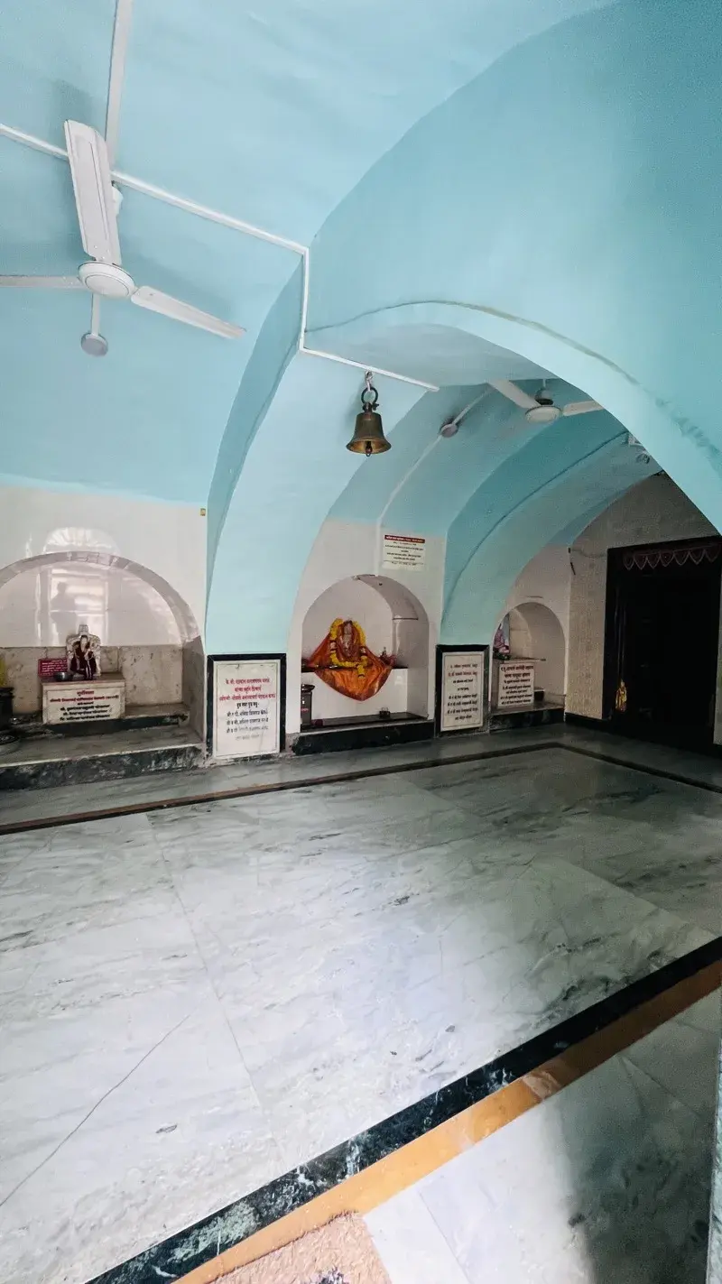 A vaulted blue corridor inside the kshetra lined with shrine niches, one holding a garlanded murti draped in saffron cloth, a bell hanging from the ceiling.