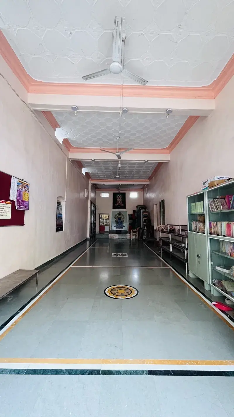 The Mahavir Sabhagruh, a long hall with ceiling fans and bookshelves along its wall, a portrait of Bhagwan Mahavir hung at the far end.